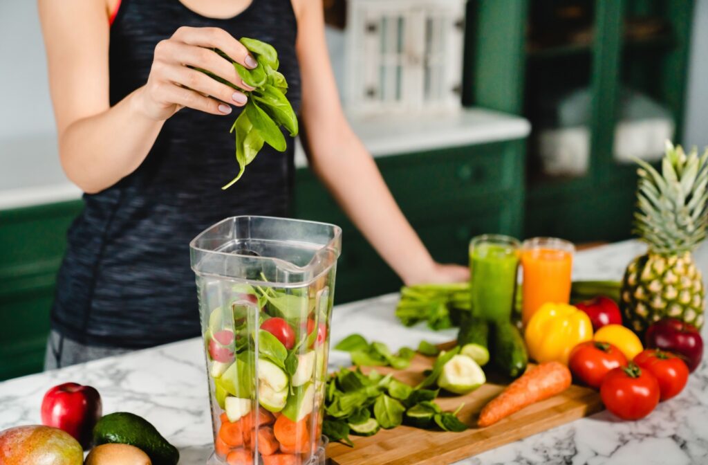 A person is holding spinach above a blender full of vegetables on a table that is also filled with vegetables